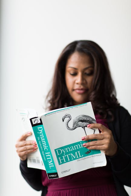 A woman with dark, wavy hair wearing a black blazer over a dark red top is holding an open book titled 'Dynamic HTML' by O'Reilly, with an illustration of a flamingo on the cover. She is standing indo