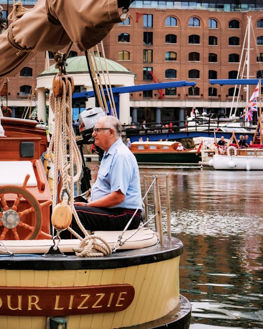 A row of sailboats moored along the edge of a calm waterway, with colorful flags fluttering from their masts. The boats are docked near a lush, green tree that provides a vibrant contrast to the surro